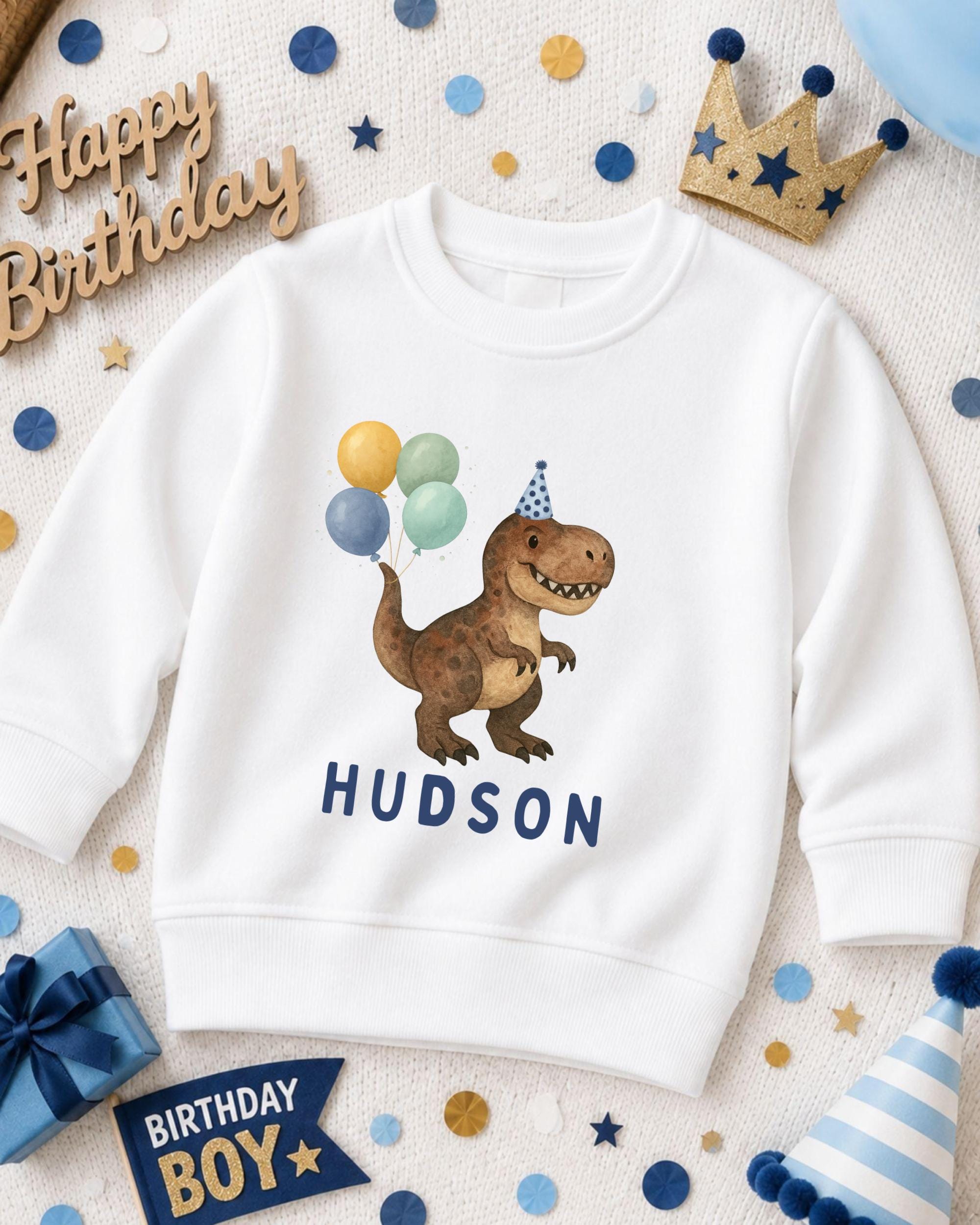 A white sweatshirt with a dinosaur wearing a party hat and balloons, surrounded by birthday decorations and a birthday banner.
