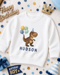 A white sweatshirt with a dinosaur wearing a party hat and balloons, surrounded by birthday decorations and a birthday banner.