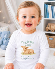 A smiling baby boy wearing a white onesie with a graphic of a cow and the text "New to the Crew" and "Peyton" printed on it.