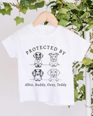 A white t-shirt with a graphic design featuring four cartoon dog illustrations and the text "PROTECTED BY Albie, Buddy, Ozzy, Teddy".