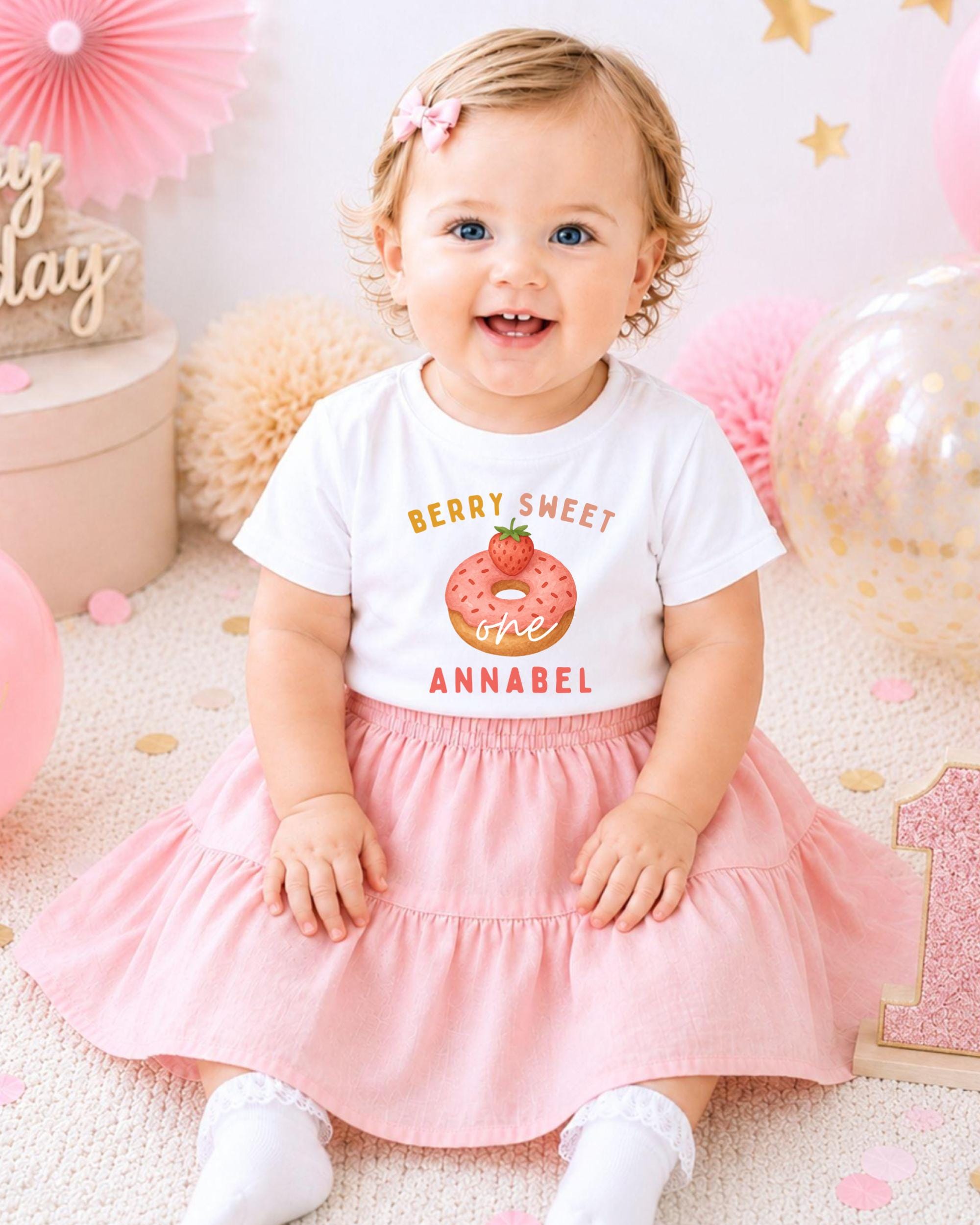 A smiling baby girl wearing a white t-shirt with a strawberry graphic and pink skirt, sitting on a pink and white background with balloons and confetti.
