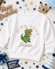A white sweatshirt with a dinosaur graphic and the text "DINO FOUR JACKSON" printed on it, surrounded by birthday decorations such as a crown, confetti, and a birthday banner.