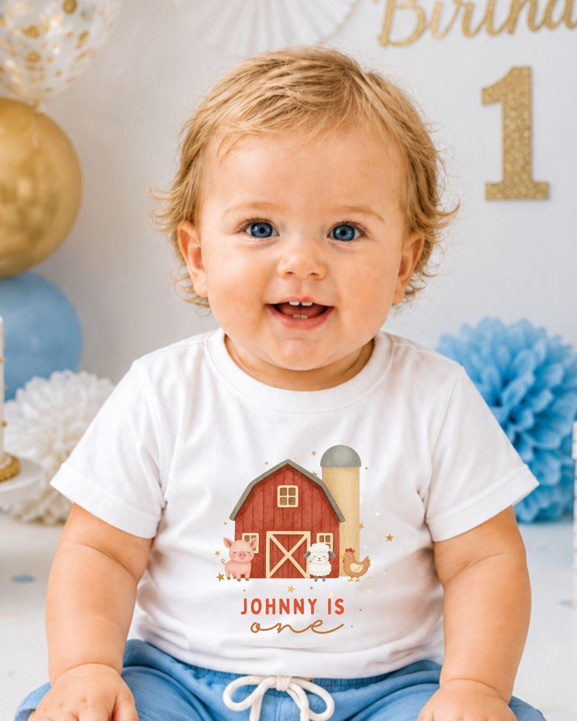 A smiling baby boy wearing a white t-shirt with a farmhouse graphic and the text "Johny is one" is sitting in front of a birthday party backdrop.