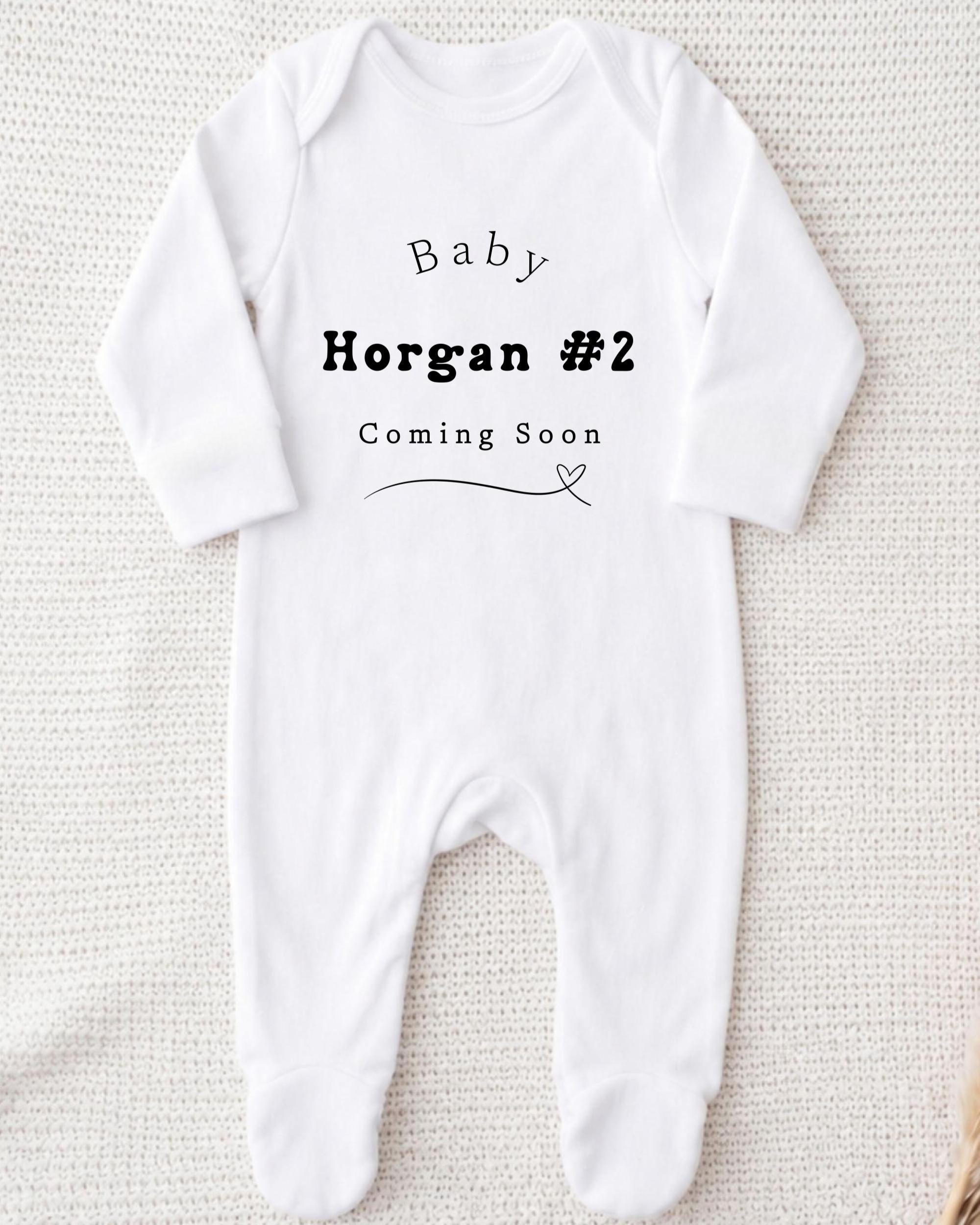 A white onesie with text printed on it, indicating that the baby is expected to arrive soon.