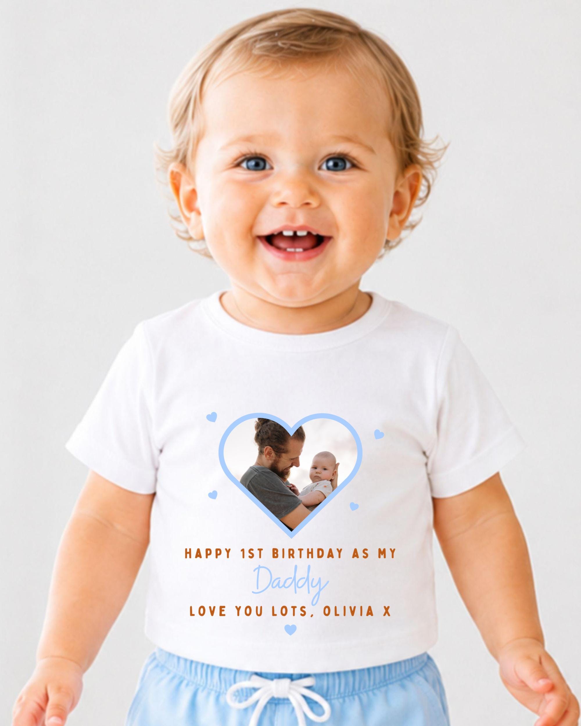 A smiling baby wearing a white t-shirt with a heart-shaped photo of a man holding a baby, along with text that reads "Happy 1st Birthday as my Daddy Love you lots, Olivia X".