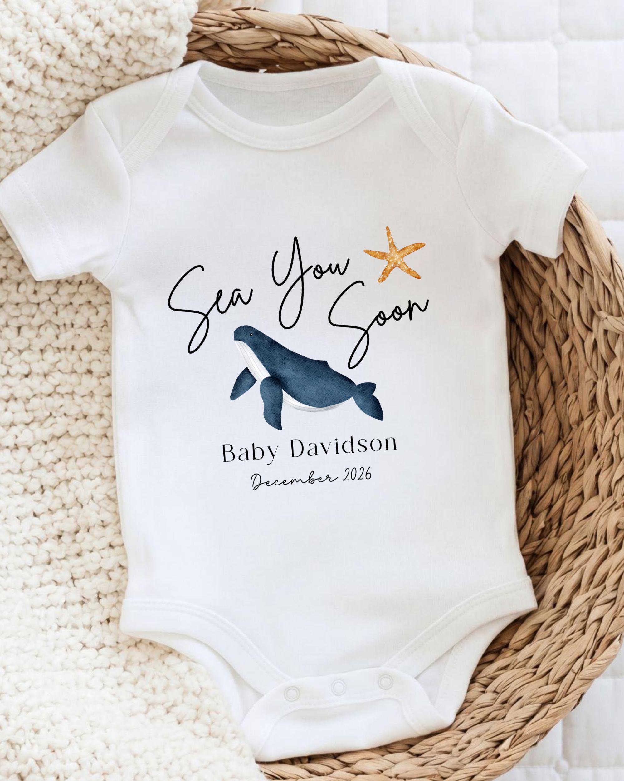 A white baby onesie with a graphic of a whale and a starfish, along with the text "Sea You Soon" and "Baby Davidson December 2026".