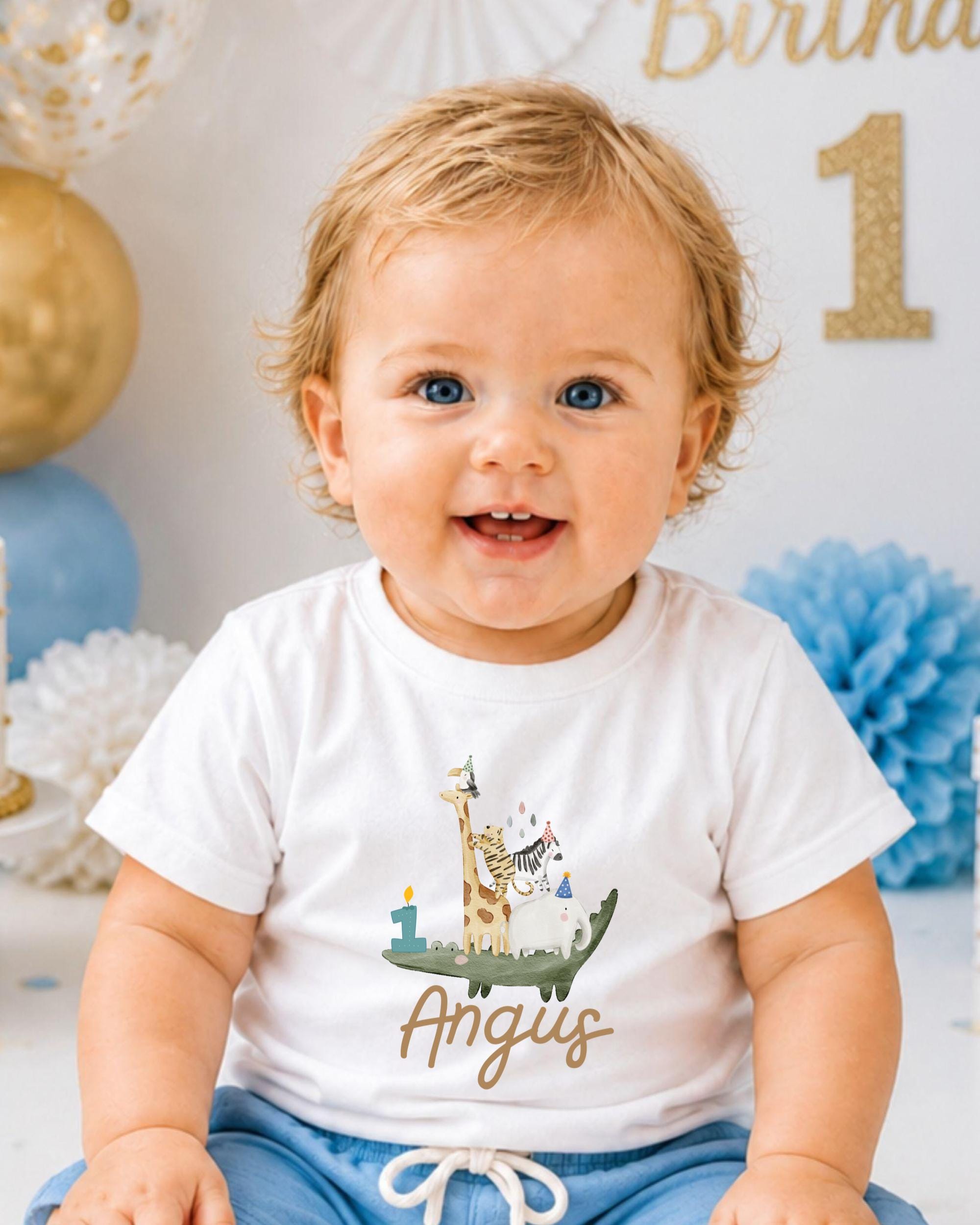 A smiling baby boy wearing a white t-shirt with a giraffe and other animal designs, sitting in front of a birthday party backdrop.