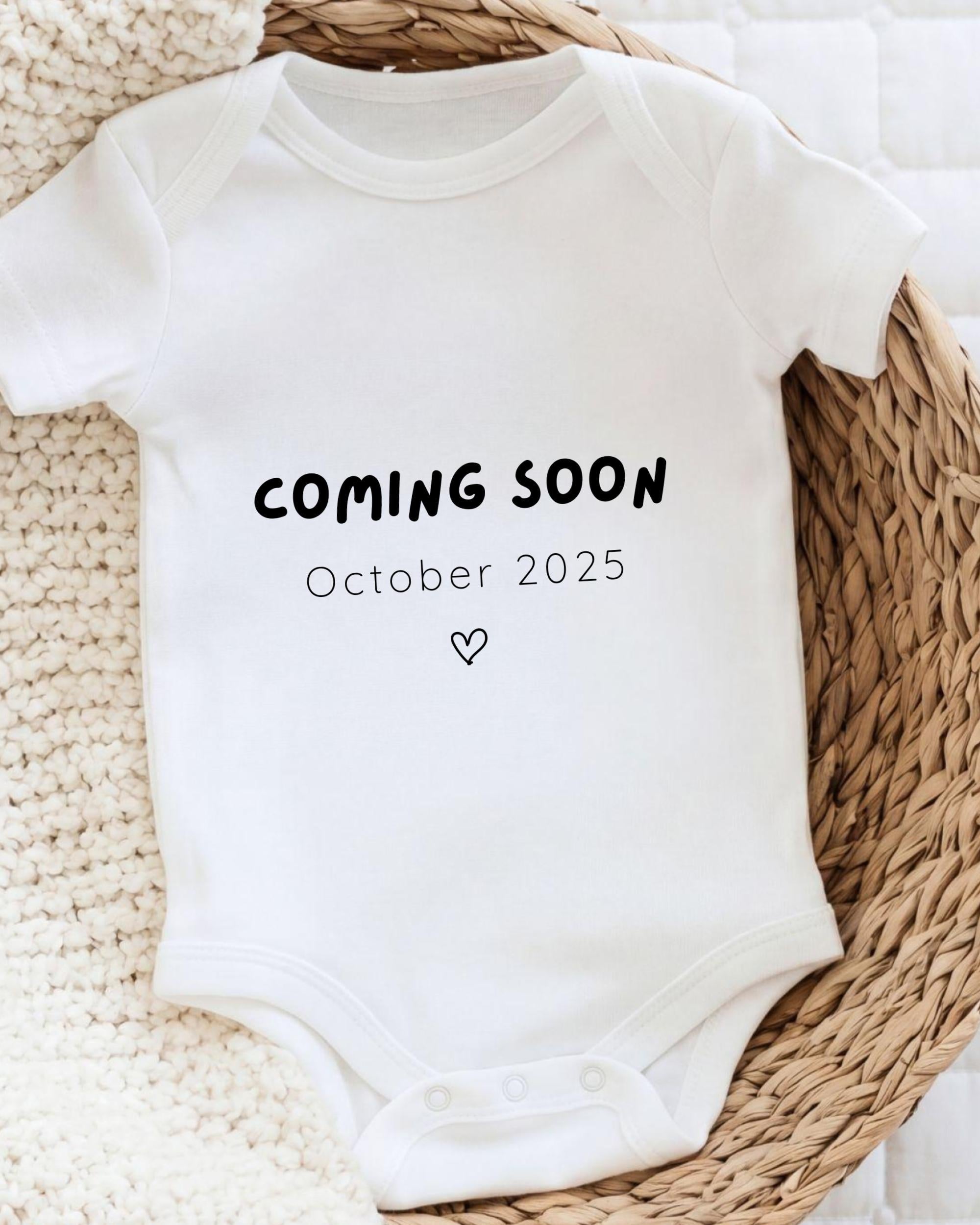 A white onesie with the text "COMING SOON October 2025" printed on it, placed on a woven basket.
