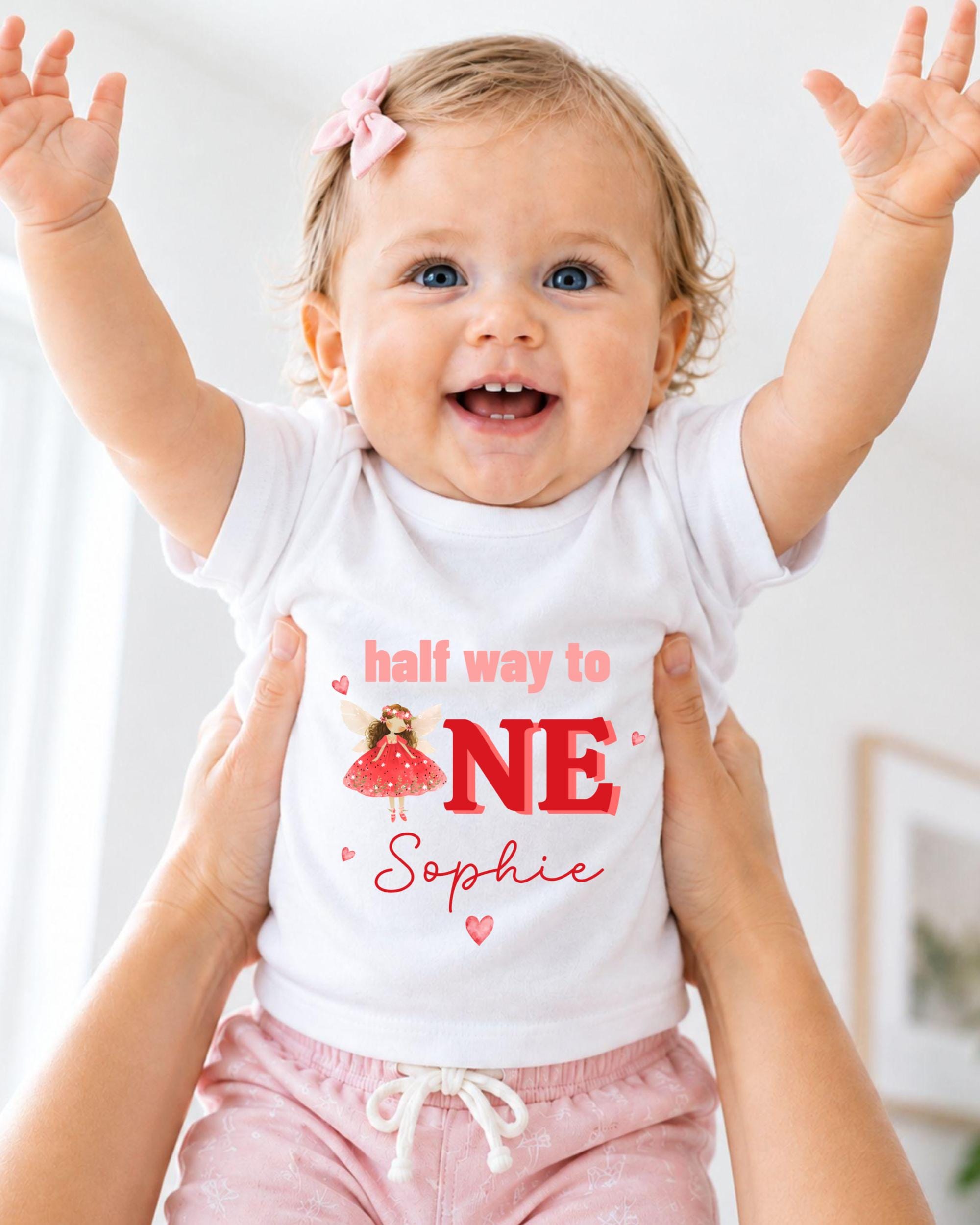 A smiling baby girl wearing a white t-shirt with the text "half way to NE Sophie" and a red character on it, being held up by someone's arms.