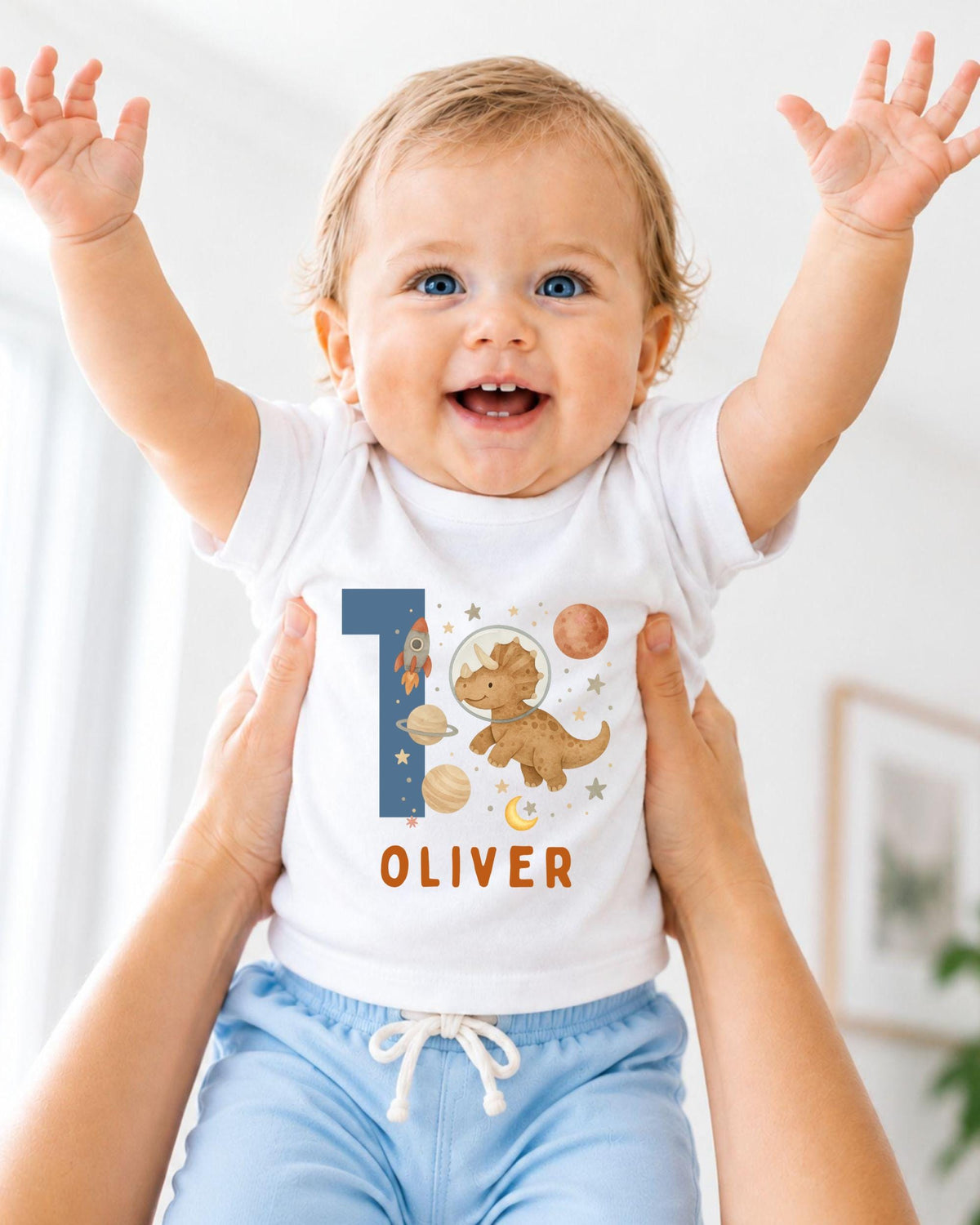 A smiling baby wearing a white onesie with a dinosaur and space-themed design, with the name "Olivier" printed on it.