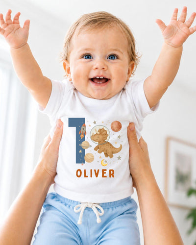 A smiling baby wearing a white bodysuit with a dinosaur and space-themed design, with the name "Olivier" printed on it.
