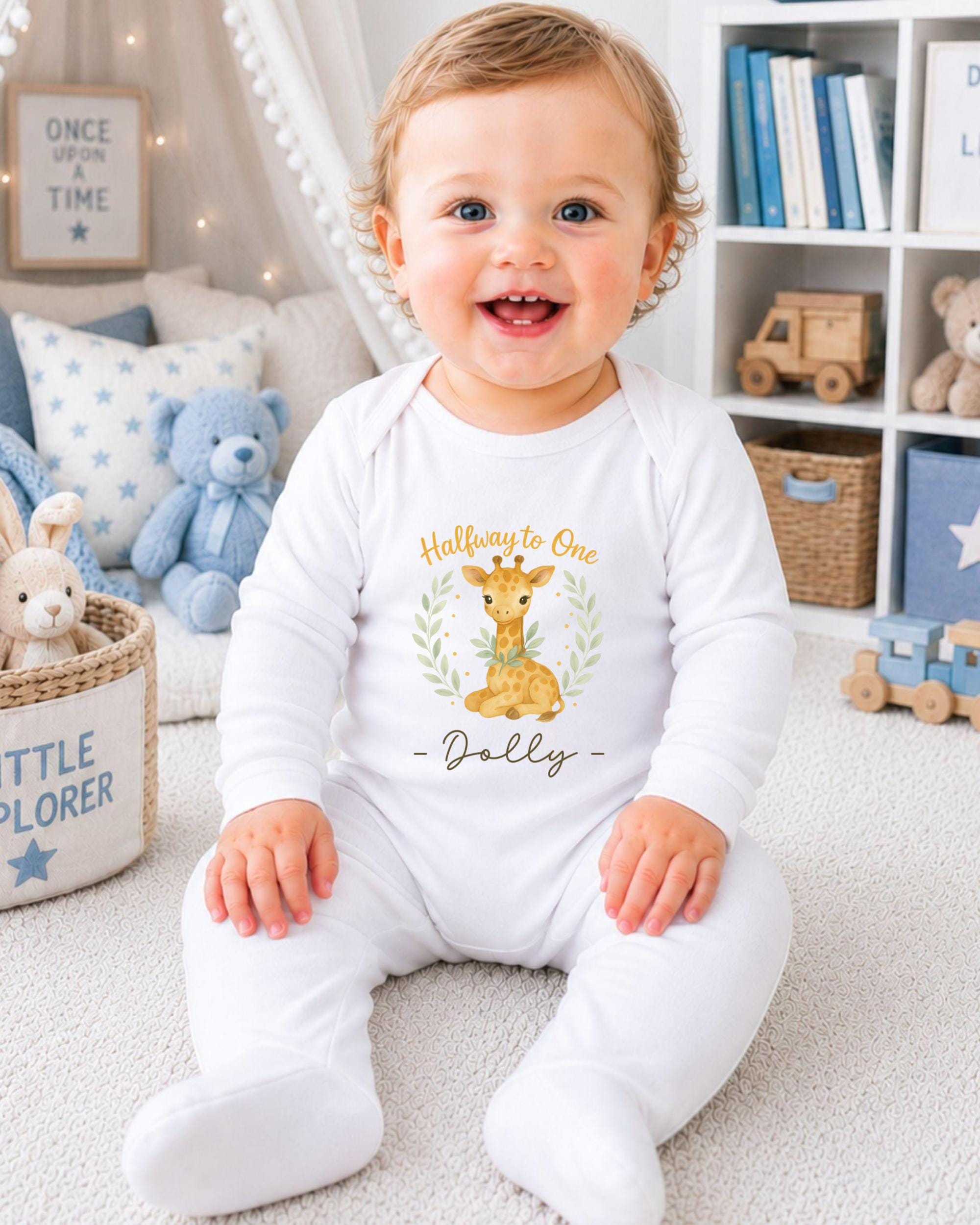 A smiling baby boy wearing a white onesie with a giraffe design and the text "Halleway to One Dolly" is sitting on the floor in a nursery setting.