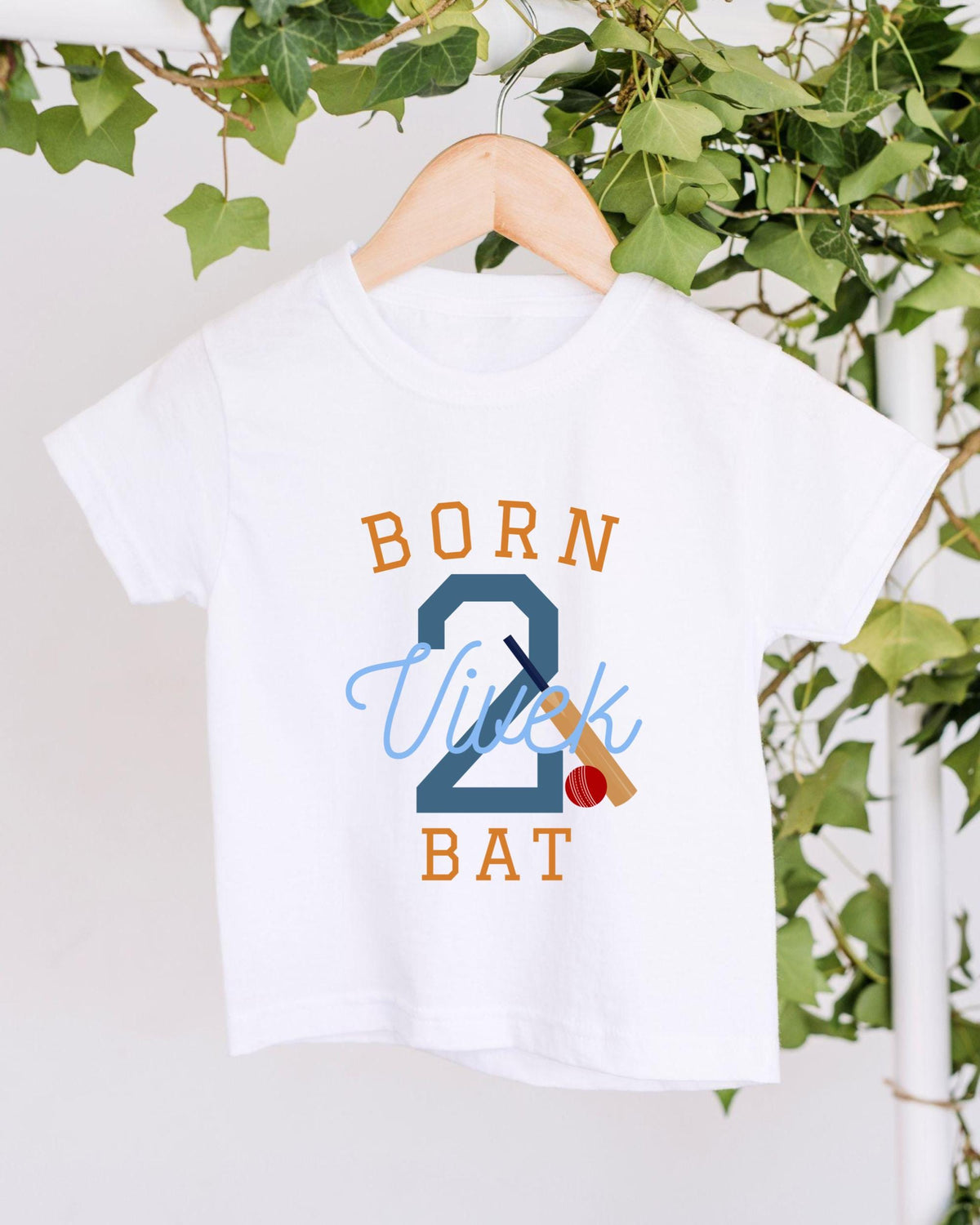 A white t-shirt with the text "BORN 2 VUITK BAT" printed on it, along with a cricket bat and ball graphic.