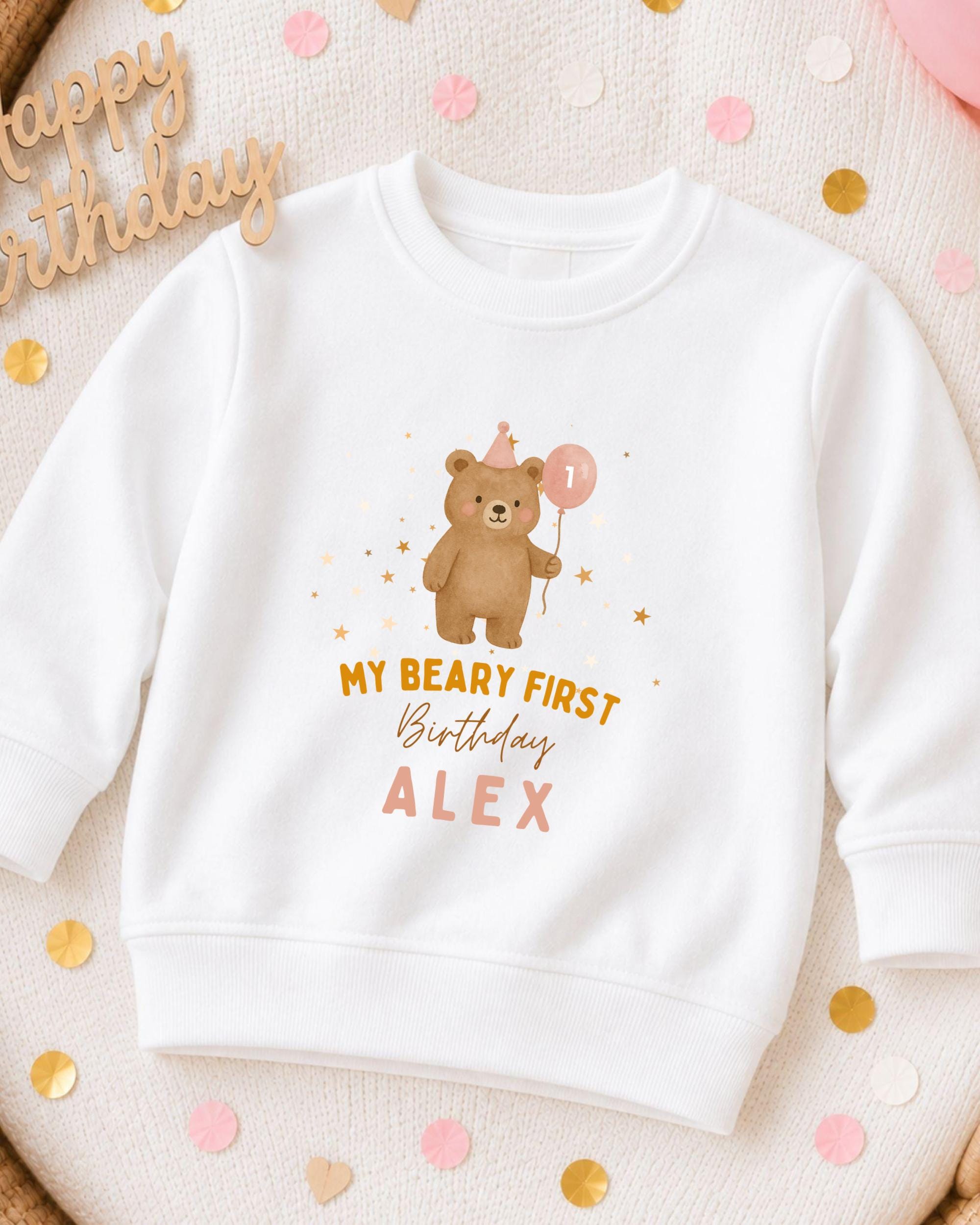 A white sweatshirt with a cartoon bear and the text "My Beary First Birthday Alex" printed on it, along with a birthday balloon.