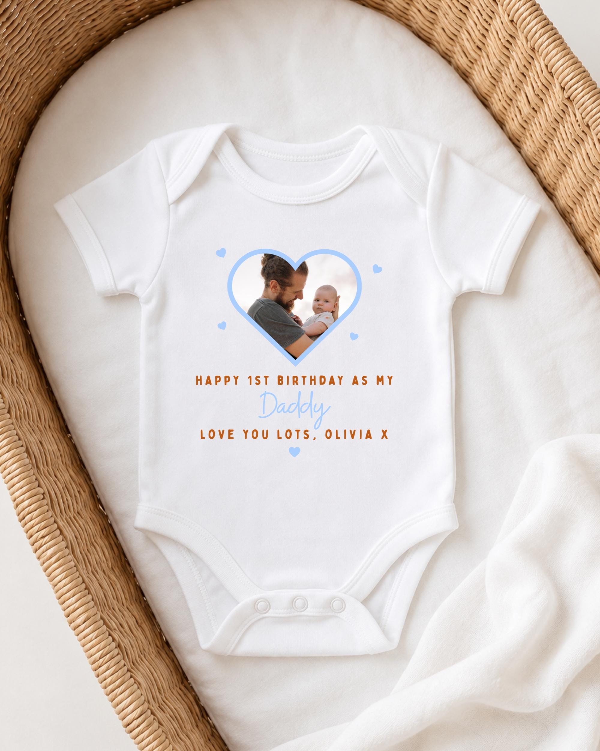 A white onesie with a photo of a man holding a baby, placed on a white blanket inside a woven basket.