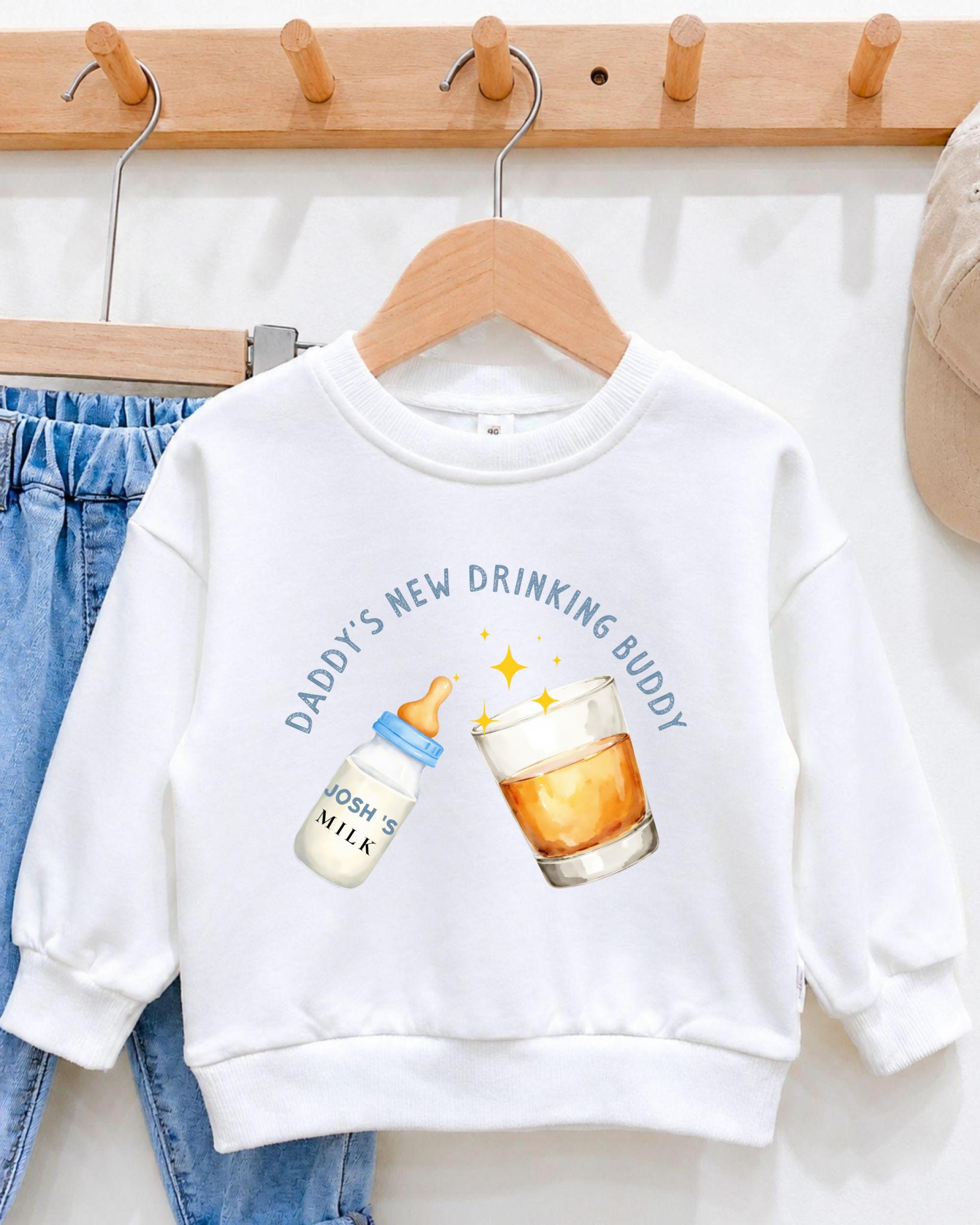 A white sweatshirt with a graphic design on it, featuring a bottle of baby milk and a glass of orange juice, along with the text "DADMY'S NEW DRINKING BUDDY".