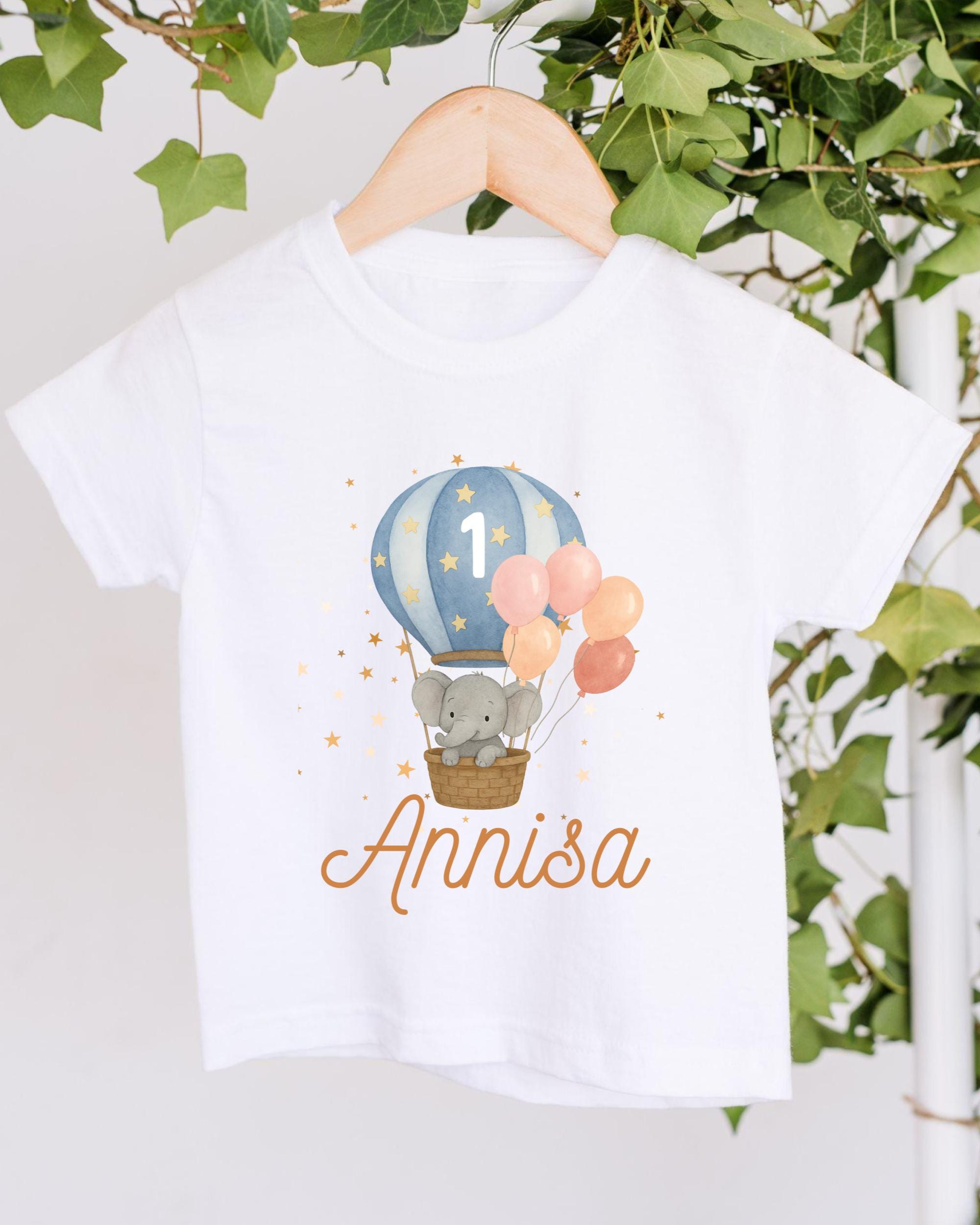 A white t-shirt with a printed design of a gray elephant in a basket, surrounded by colorful balloons, and the name "Annisa" written below.