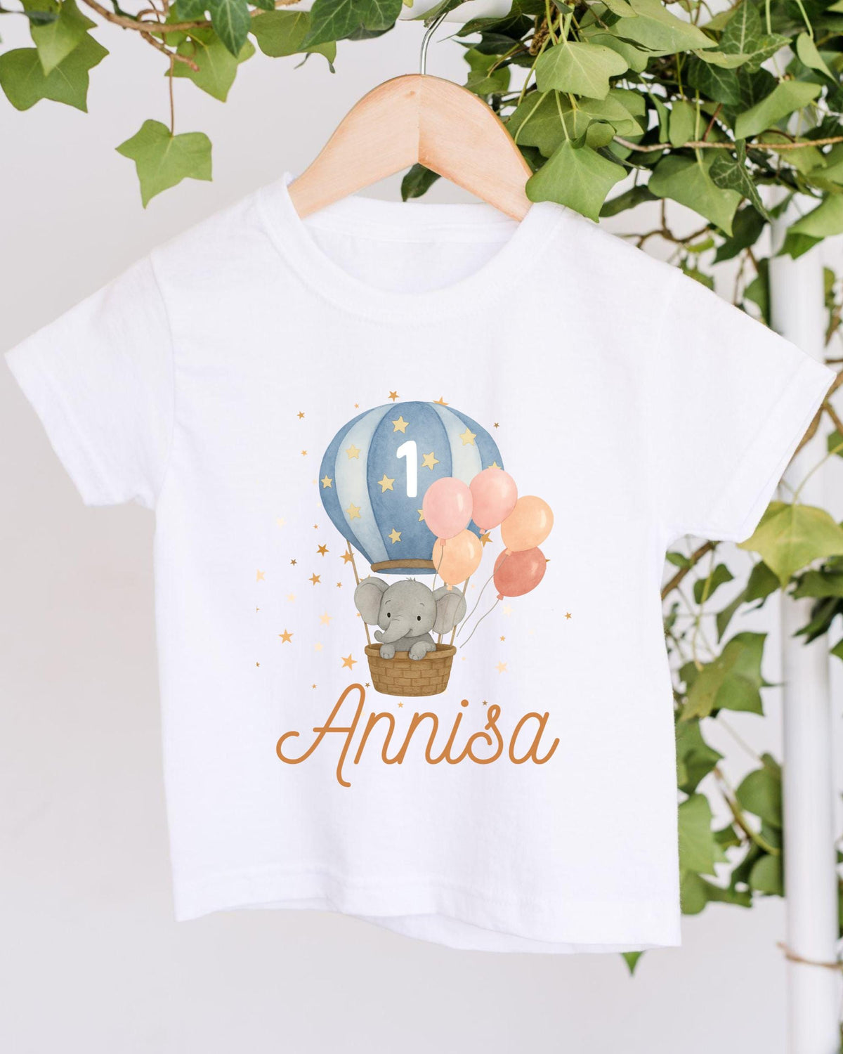 A white t-shirt with a printed design of a gray elephant in a basket, surrounded by colorful balloons, and the name "Annisa" written below.
