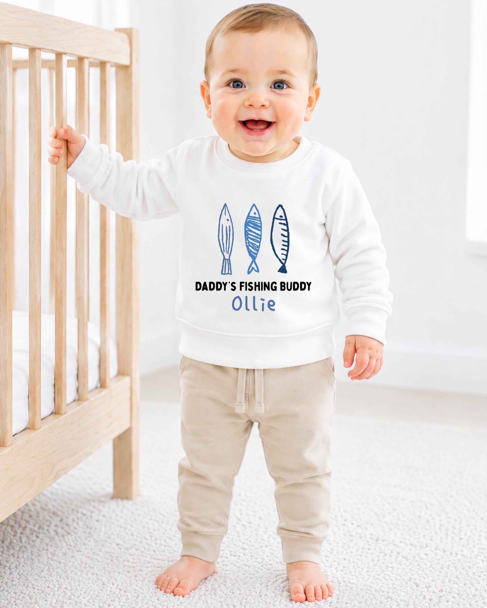 A smiling baby boy wearing a white sweatshirt with a design of fish and the text "DADDY'S FISHING BUDDY OLLIE" is standing next to a wooden crib.