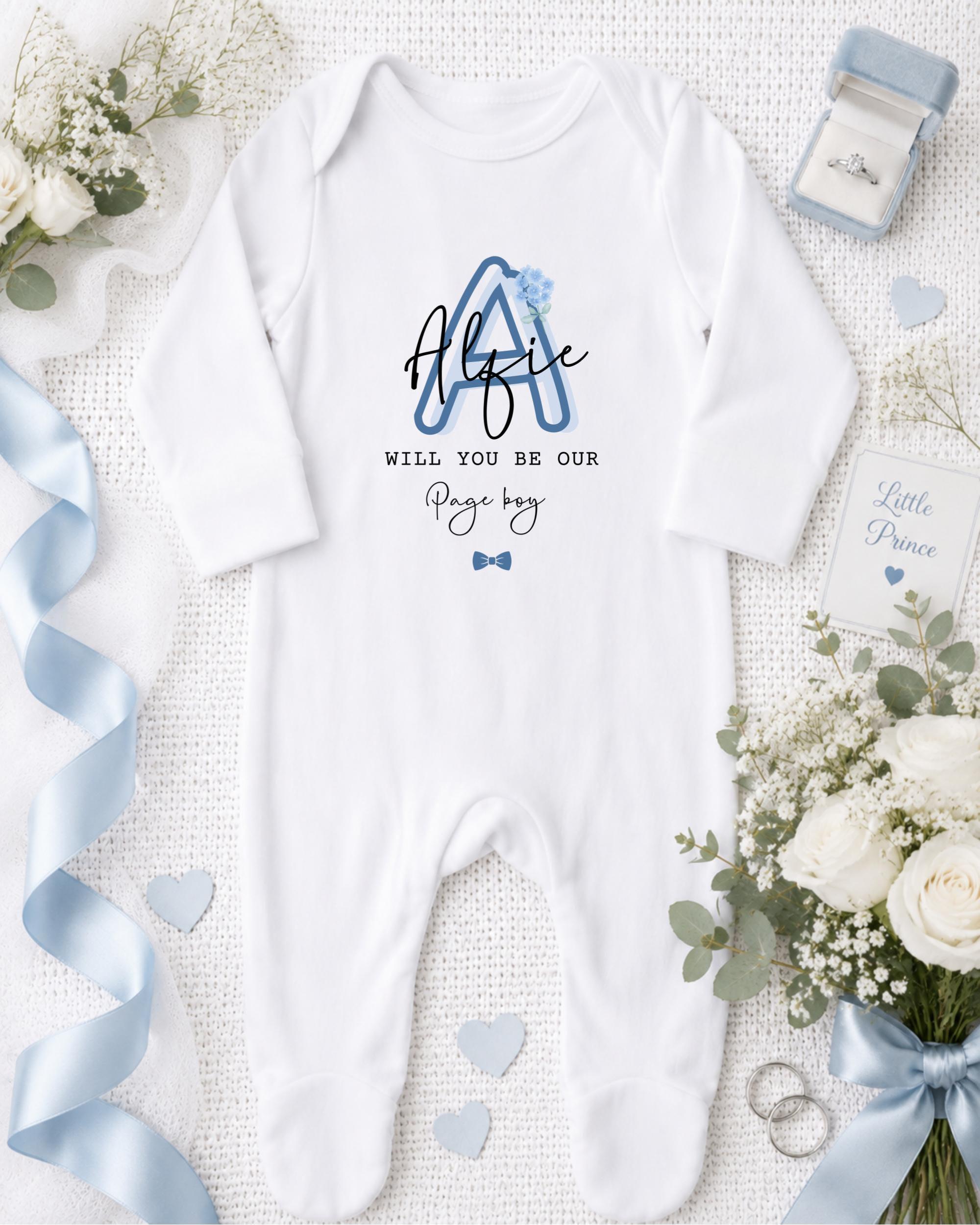 A white onesie with a blue letter "A" and the text "Will you be our Page boy?" is displayed on a bed with white flowers and a blue ribbon.