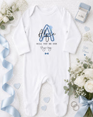 A white onesie with a blue letter "A" and the text "Will you be our Page boy?" is displayed on a bed with white flowers and a blue ribbon.