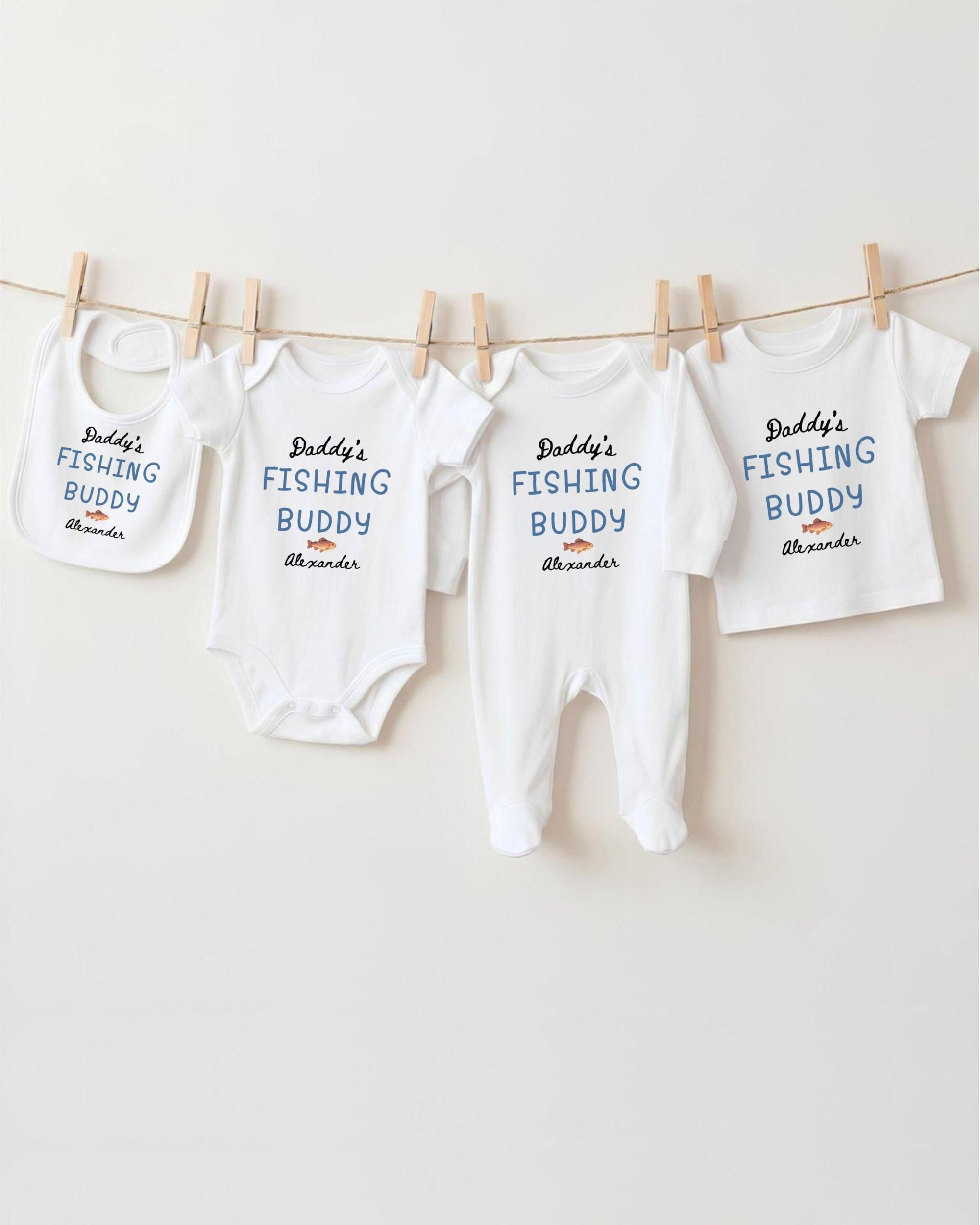 A row of white baby clothes with the text "Daddys Fishing Buddy Alexander" printed on them, hanging on a clothesline.