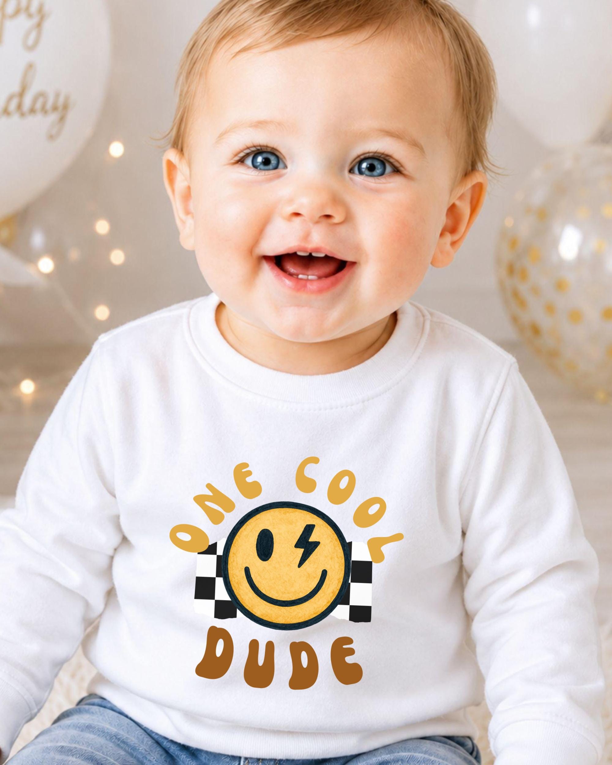 A smiling baby boy wearing a white shirt with a smiley face and the text "One cool dude" printed on it.
