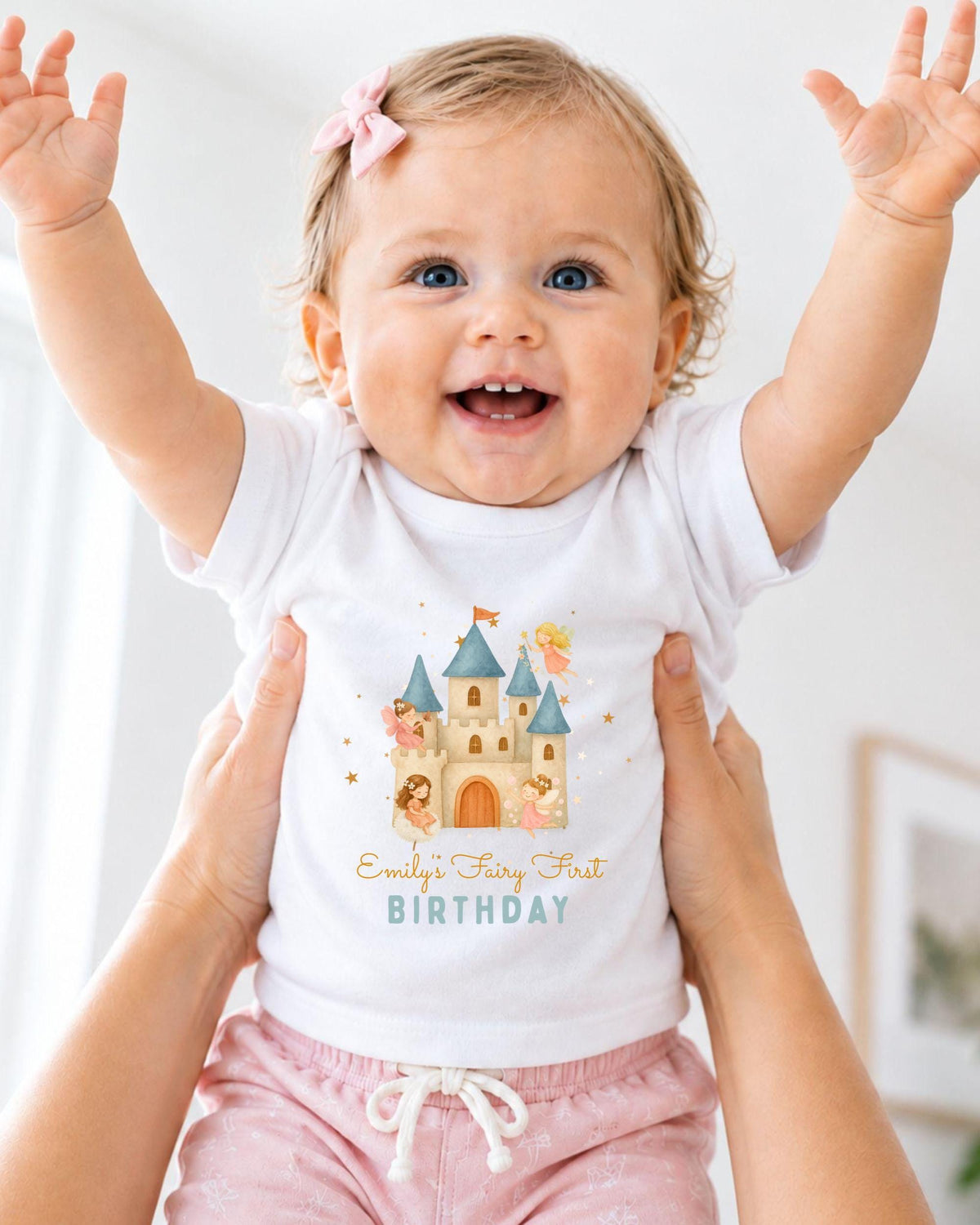 A smiling baby girl wearing a white t-shirt with a castle graphic and the text "Emilly's Fairy First Birthday" is being held up by someone's arms.