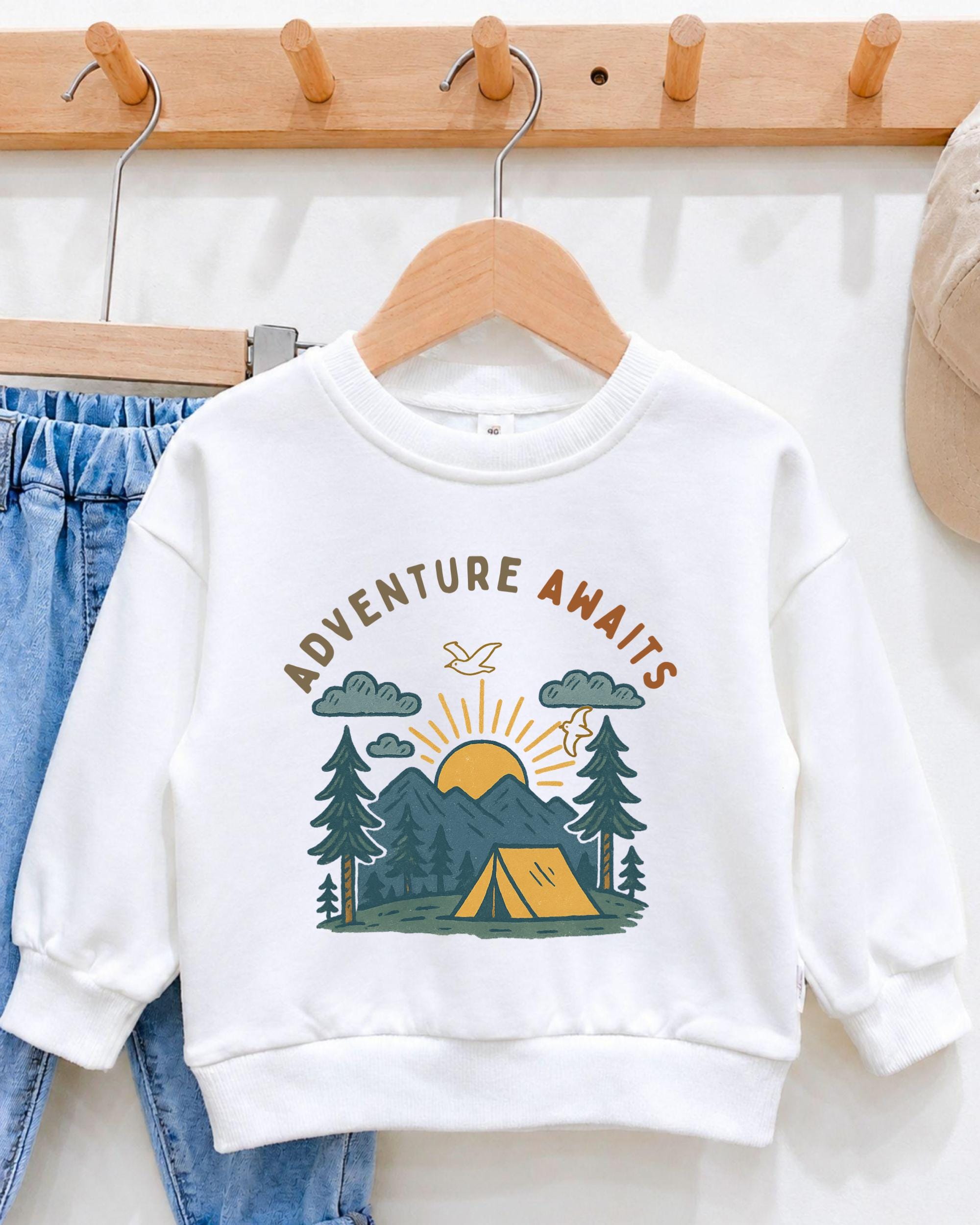 A white sweatshirt with a graphic design of a camping scene and the text "Adventure Awaits" printed on it, hanging on a wooden hanger.