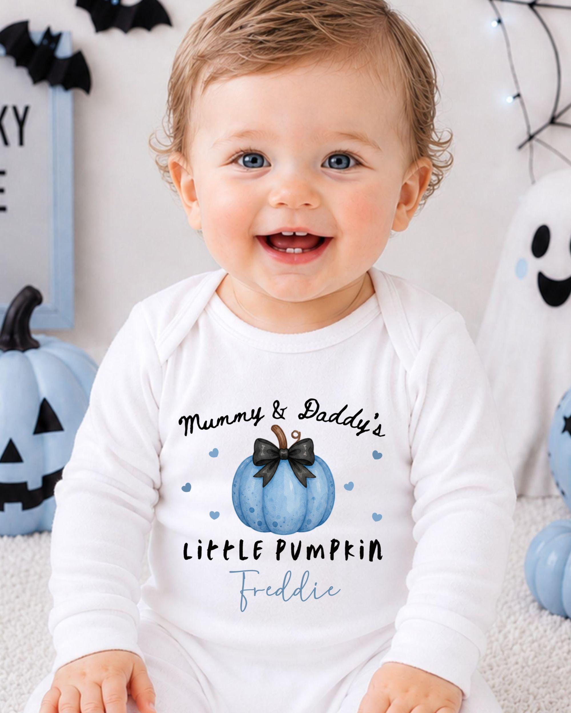 A smiling baby boy wearing a white onesie with a pumpkin and text on it, sitting in front of Halloween decorations.