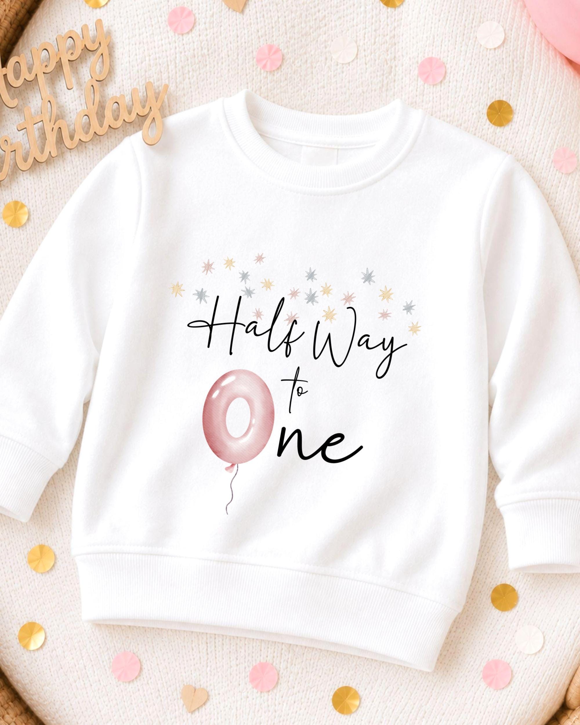 A white sweatshirt with a graphic design on it, featuring a pink balloon and the text "Half Way to One". The sweatshirt is placed on a surface with confetti and a wooden sign that says "Happy Birthday".