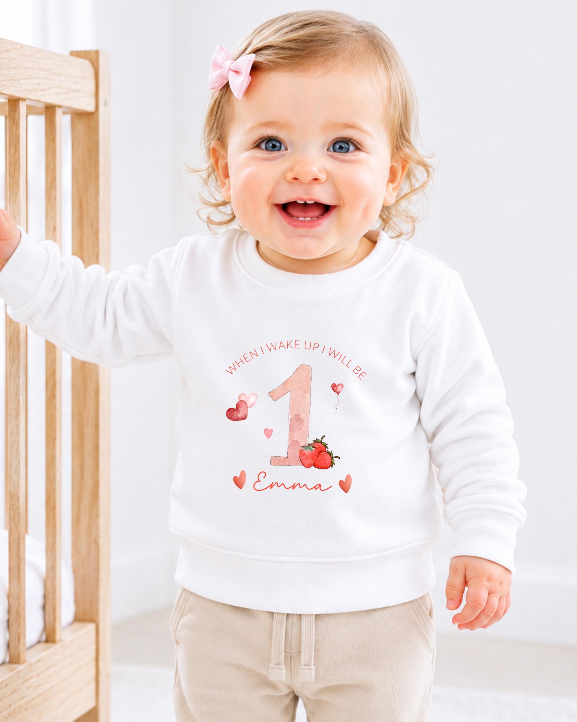 A smiling baby girl wearing a white sweater with a personalized design and text.