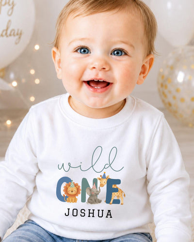 A smiling baby boy wearing a white long-sleeved shirt with a personalized text and animal graphics.