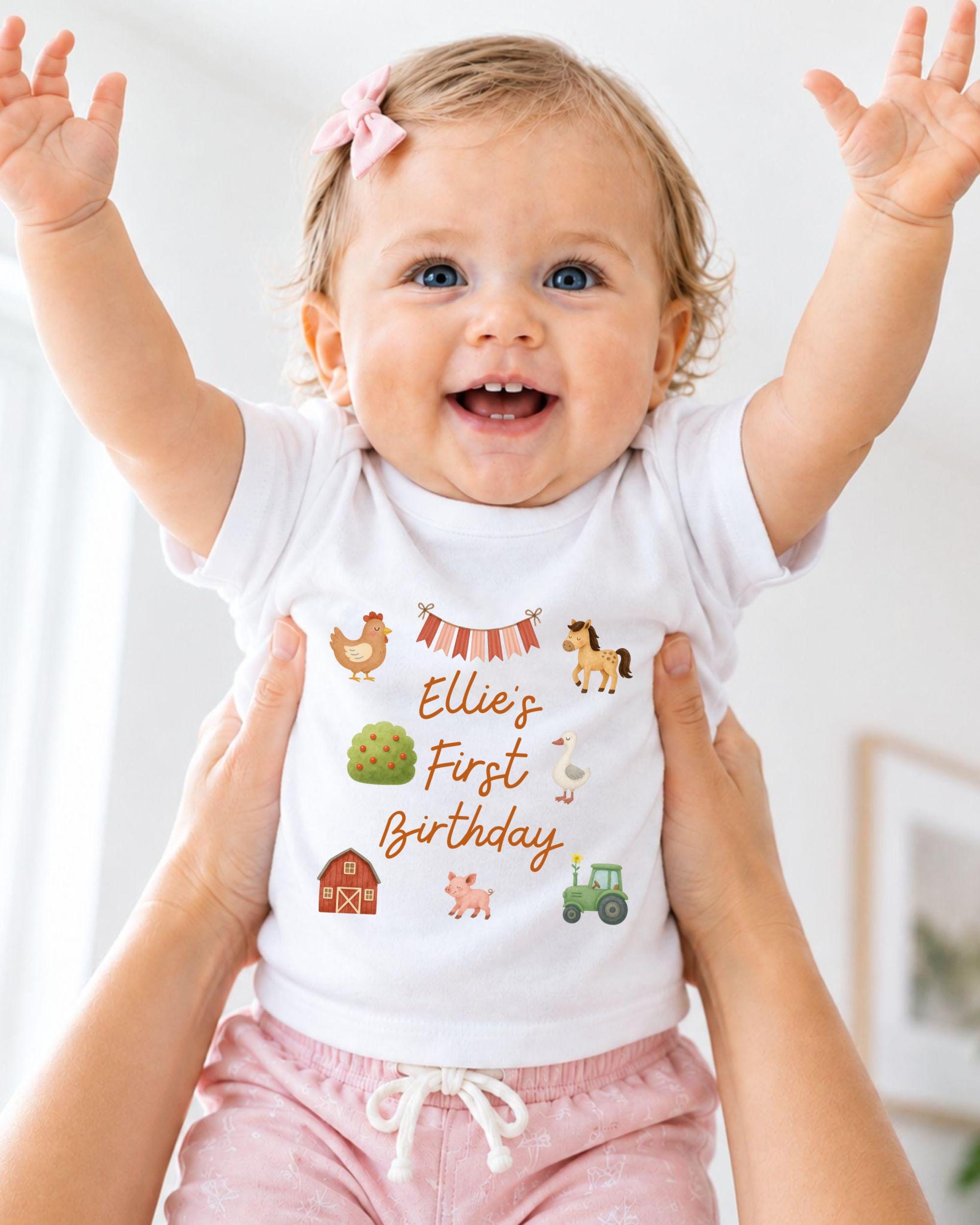 A smiling baby girl wearing a white t-shirt with farm animal illustrations and the text "Ellie's First Birthday" is being held up by someone's arms.