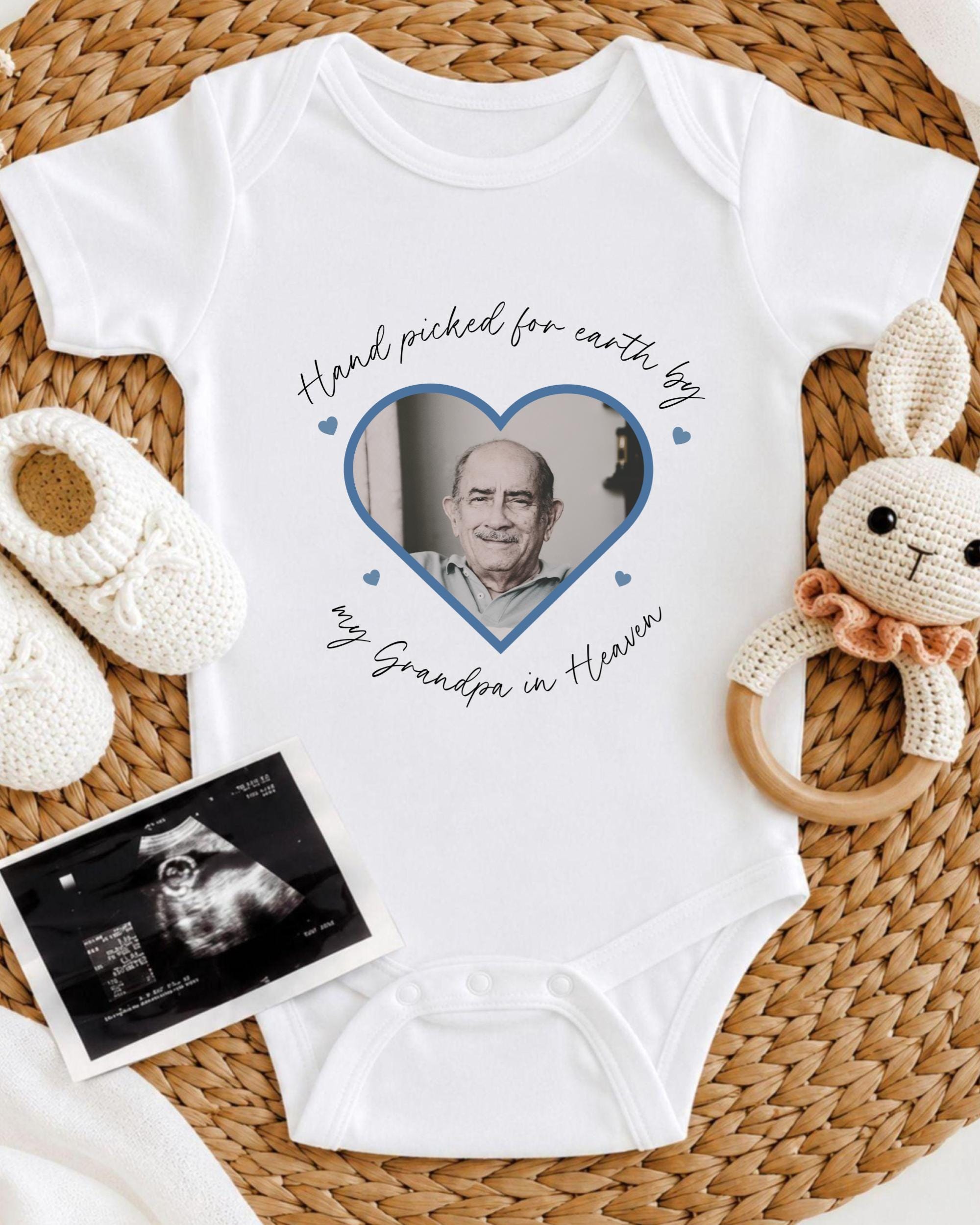 A white baby onesie with a photo of an older man inside a heart shape, surrounded by a crocheted bunny toy and a baby bootie.