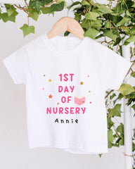 A white t-shirt with pink text and stars hanging from a wooden hanger, placed against a backdrop of green ivy leaves.