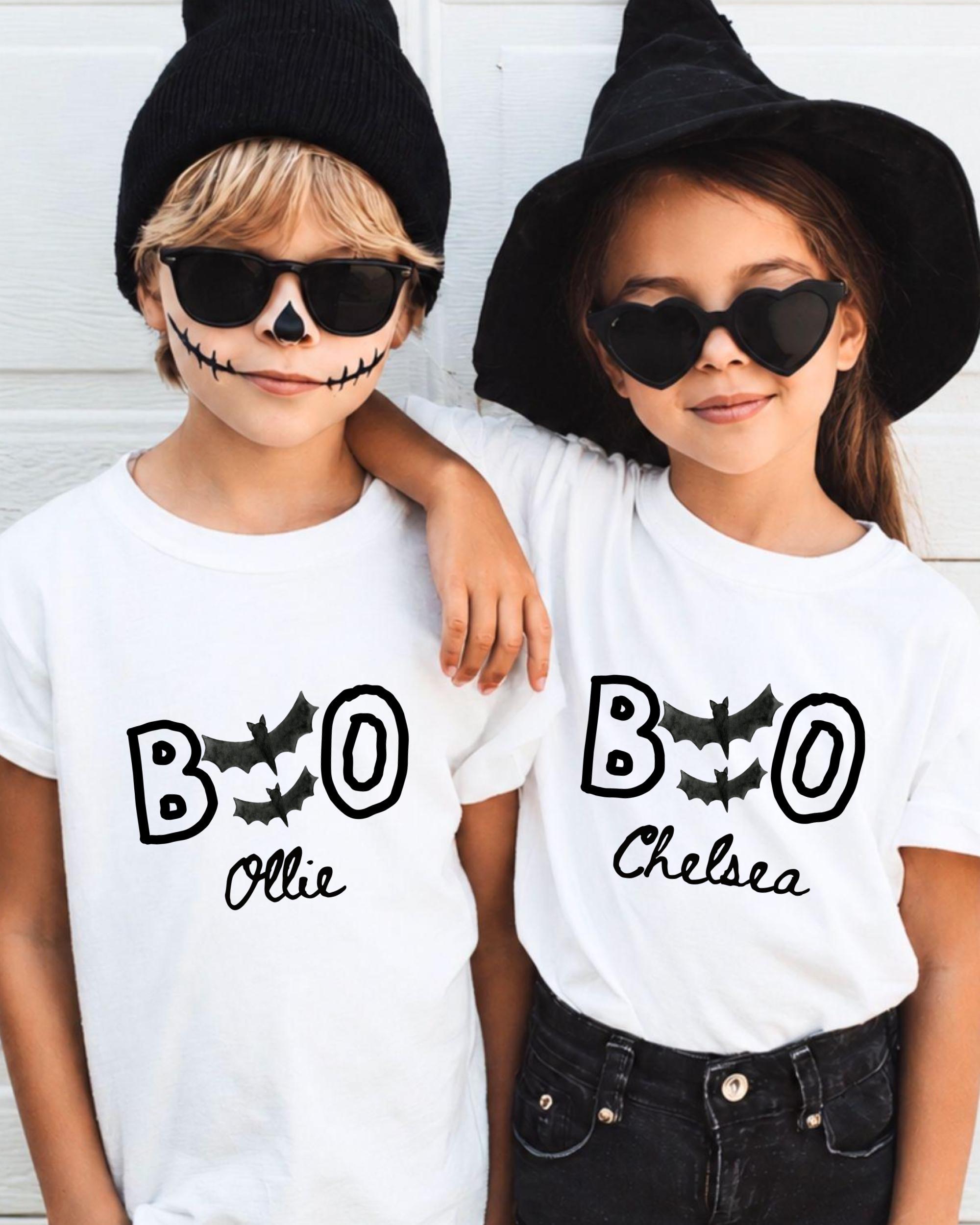 Two children wearing white t-shirts with the text "BOO Chelsea" printed on them, along with bats and a skeleton face design, posing for a photo.