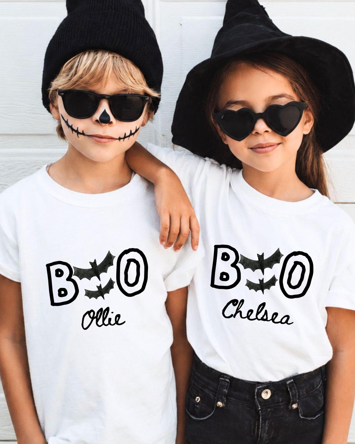 Two children wearing white t-shirts with the text "BOO Chelsea" printed on them, along with bats and a skeleton face design, posing for a photo.