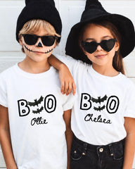 Two children wearing white t-shirts with the text "BOO Chelsea" printed on them, along with bats and a skeleton face design, posing for a photo.