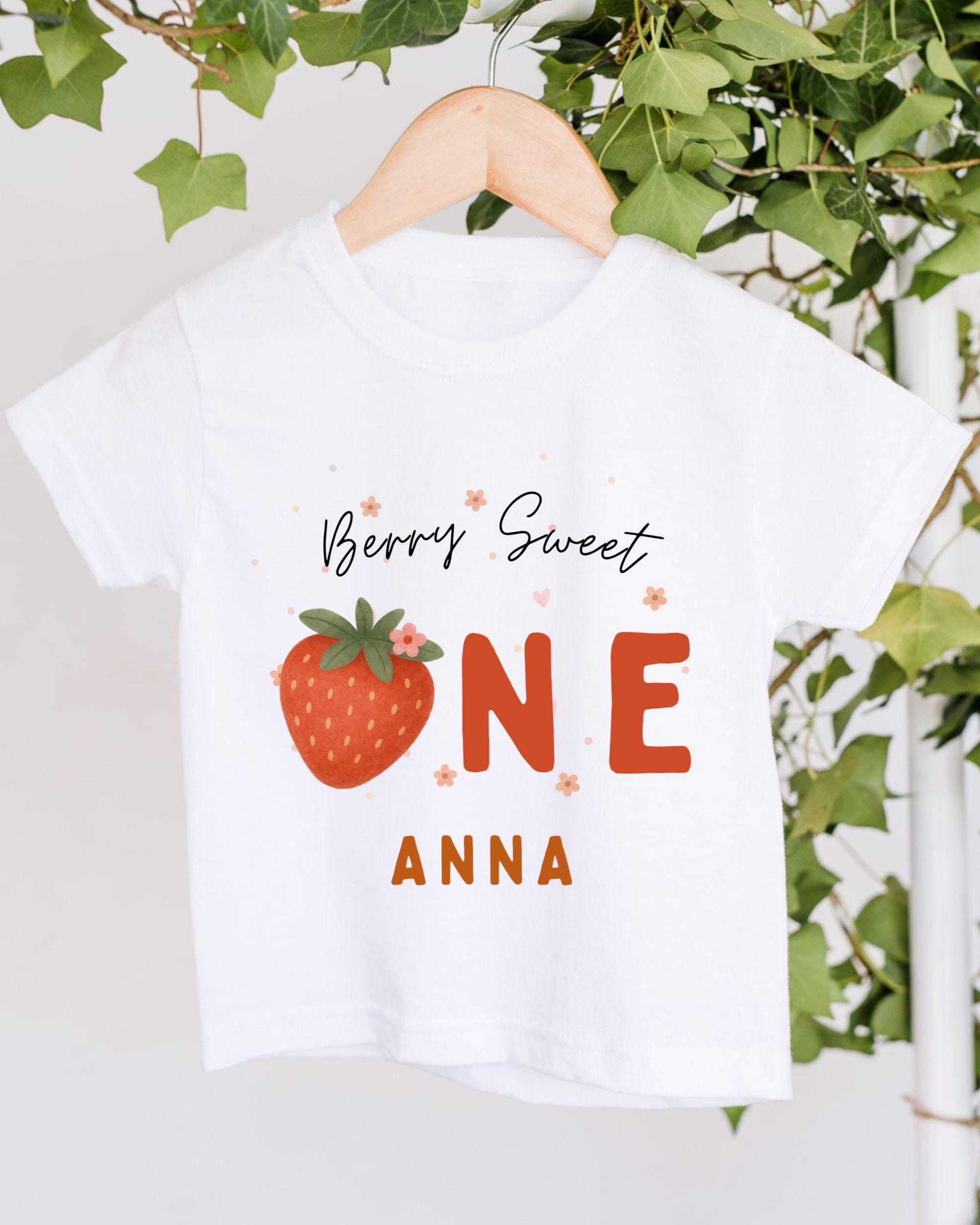 A white t-shirt with a graphic design featuring a strawberry and the text "Berry Sweet One Anna" printed on it.