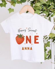 A white t-shirt with a graphic design featuring a strawberry and the text "Berry Sweet One Anna" printed on it.
