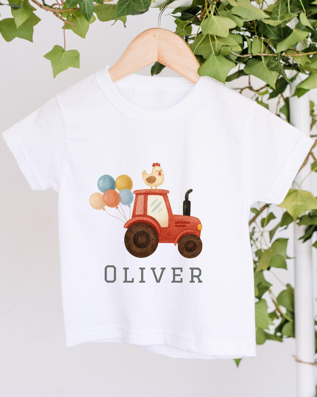A white t-shirt with a graphic design of a red tractor carrying colorful balloons and a chicken on top, along with the name "Olivier" printed below.