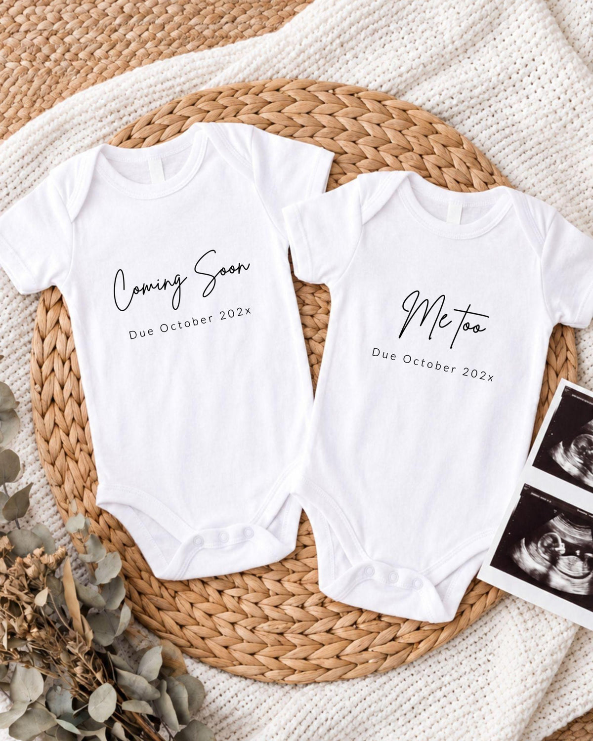 Two white baby onesies with text that announces the upcoming arrival of a baby, placed on a woven placemat.