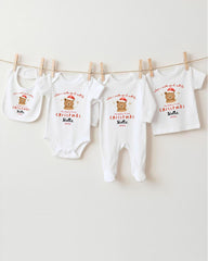 A row of four white Christmas-themed baby clothes, including onesies and bibs, hanging on a clothesline.