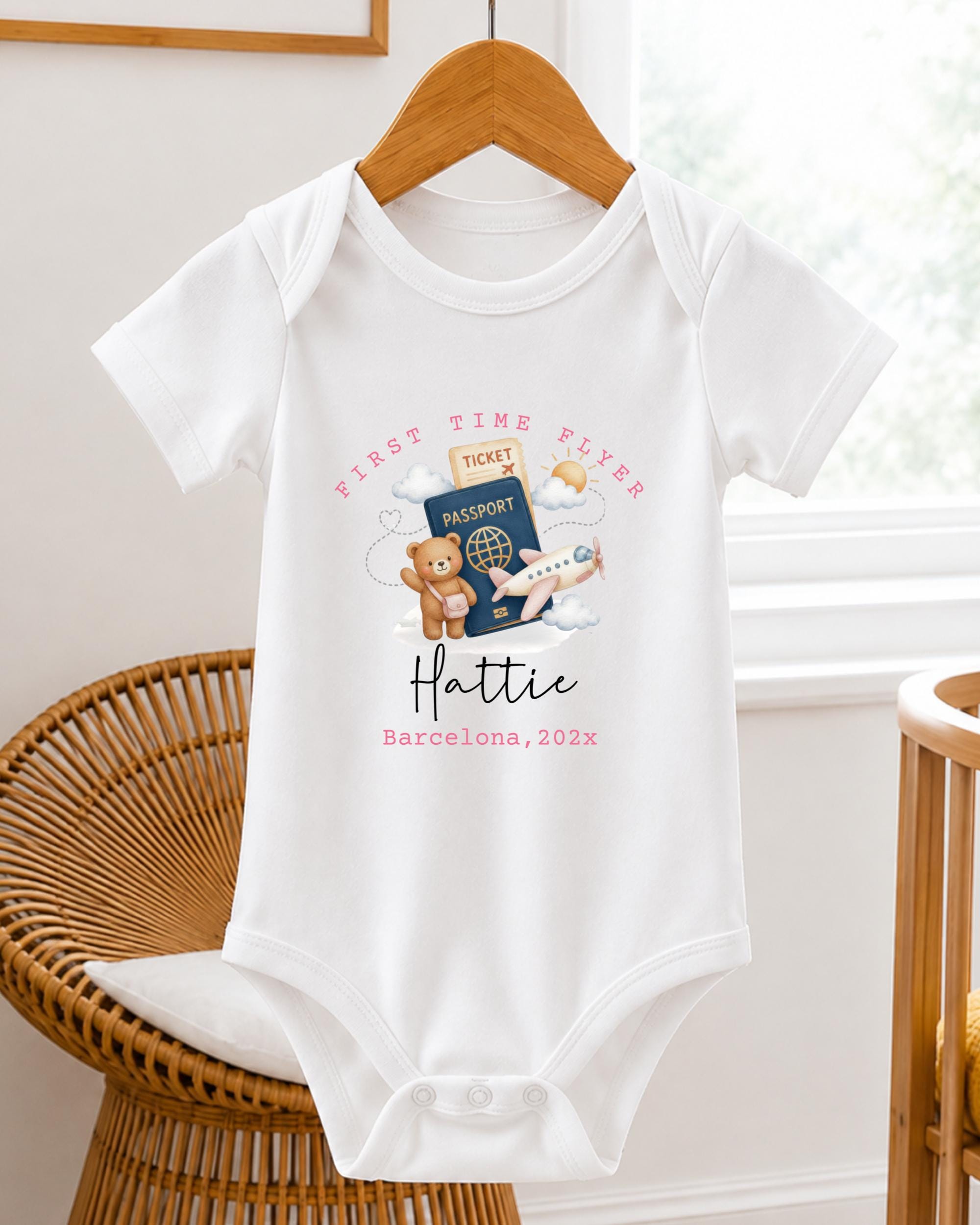 A white baby onesie with a travel-themed design, including a passport, airplane, and teddy bear, hangs on a wooden hanger.