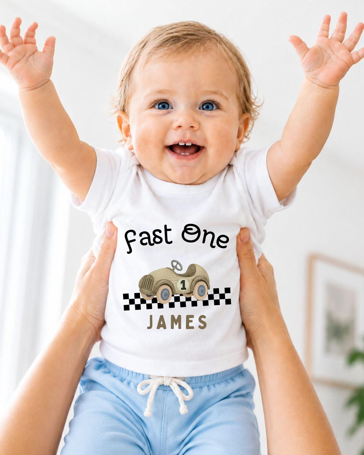 A smiling baby wearing a white t-shirt with a race car graphic and the text "Fast One James" is being held up by someone's arms.