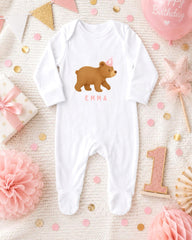A white onesie with a bear design and the name "Emma" printed on it, along with a pink and gold birthday decoration, pink and gold confetti, and a pink and gold star-shaped ornament.