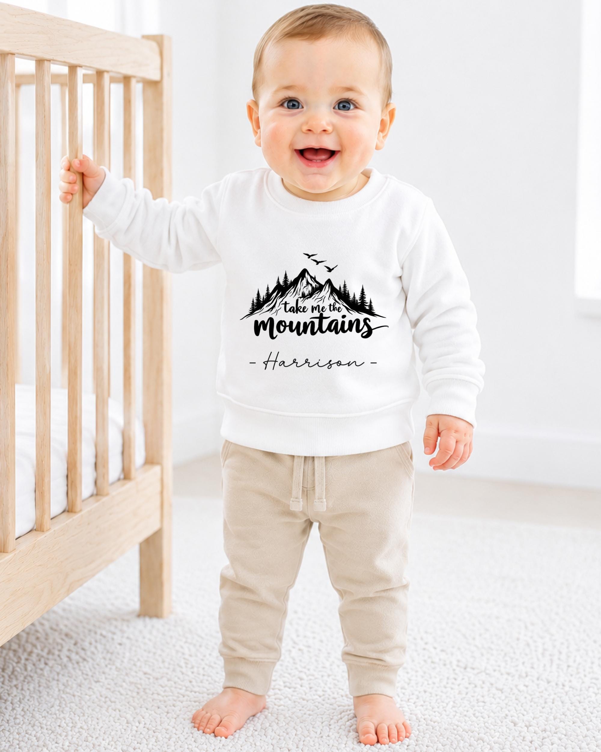 A smiling baby boy wearing a white sweatshirt with a mountain design and the text "Take a mountain" is standing next to a wooden crib.