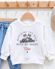 A white sweatshirt with a racing car design and the text "Watching the race with my daddy" printed on it, hanging on a wooden hanger.