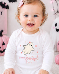 A smiling baby girl wearing a white onesie with a ghost and the text "Little Bootful Zara" is sitting in front of Halloween decorations.